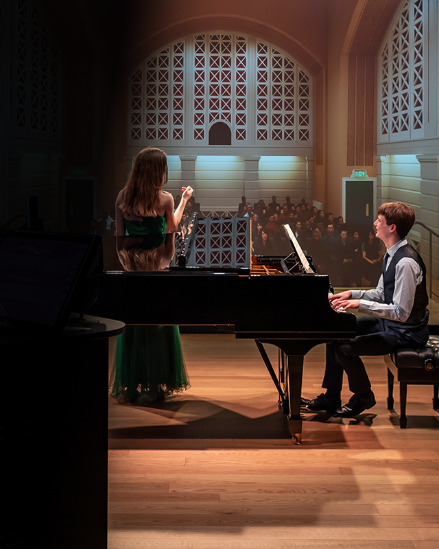 A male and female student, wearing formal attire, singing and playing the piano, in front of a performance simulator.